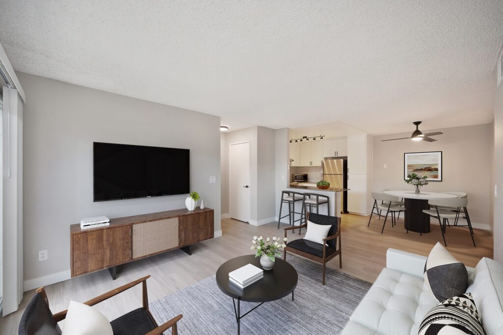 Open-concept living room with views of renovated kitchen and dining area. Space has a dresser with a television above it and two arm chairs.