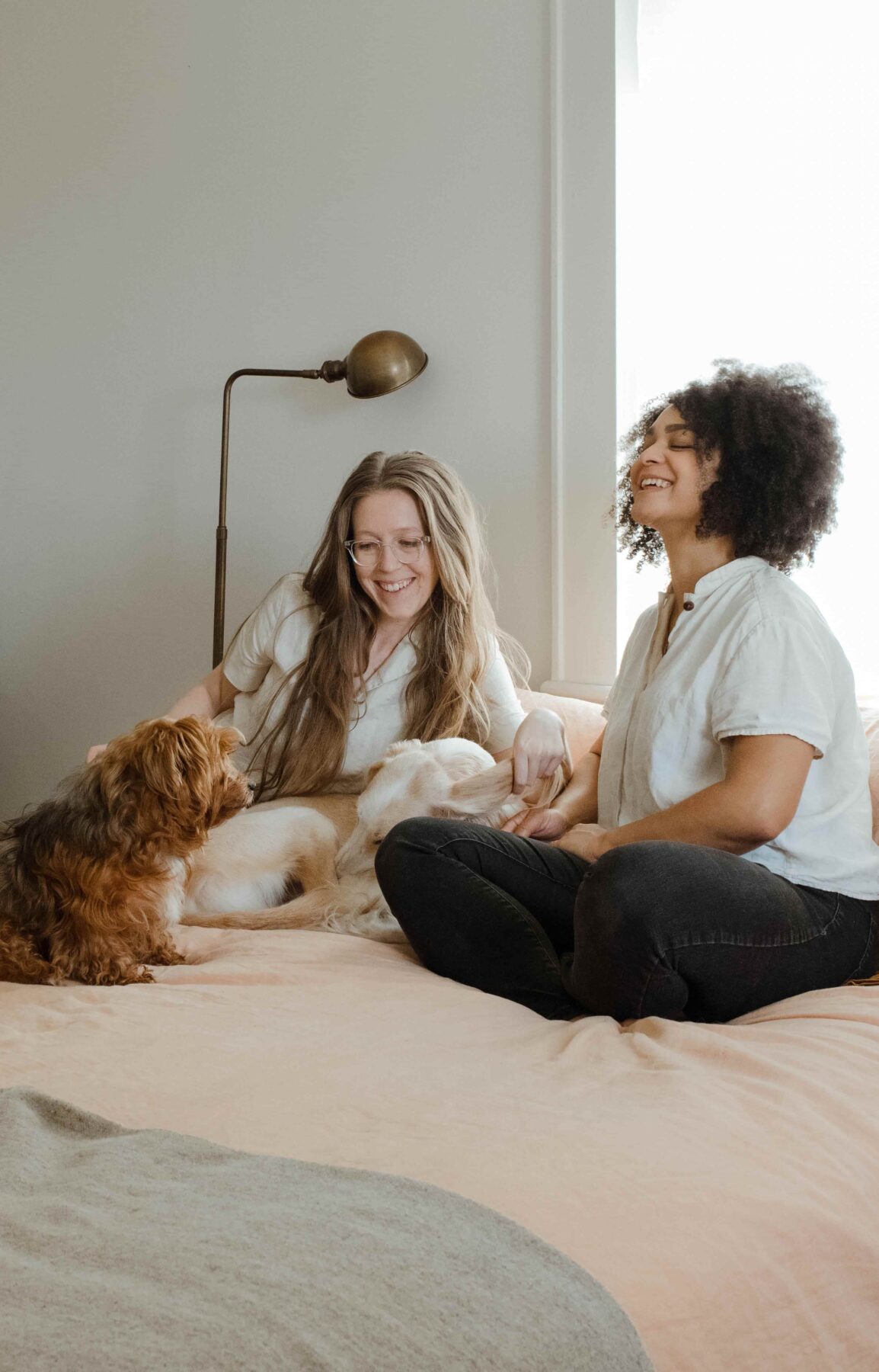 Two women sit on a bed smiling and petting two dogs. One woman has long hair and the other has curly hair. A lamp is on the wall behind them, and soft natural light comes through a window.